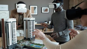 Group of architects in vr headsets standing around table with house layout during presentation of new block of flats
